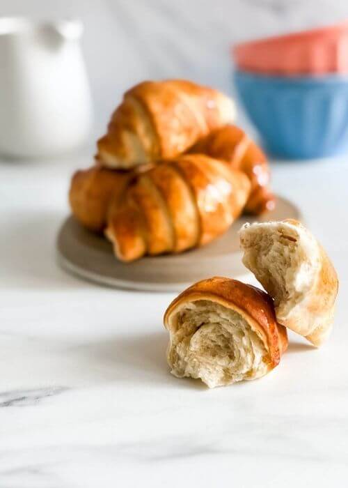 A broken croissant air fryer sits in the foreground, showing off its fluffy, layered interior, while a plate of whole, golden-brown croissants rests behind it on a beige plate.