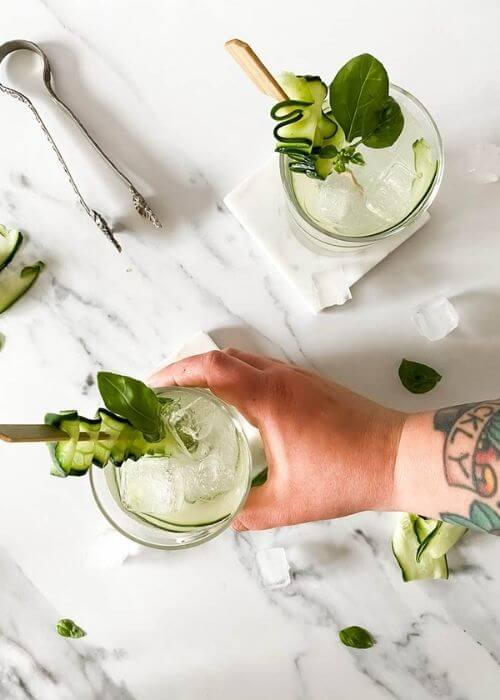 from top - two glasses of cucumber basil mocktail on marble coasters and on a marble background, around ice, basil, cucumber slices and an ice tongs