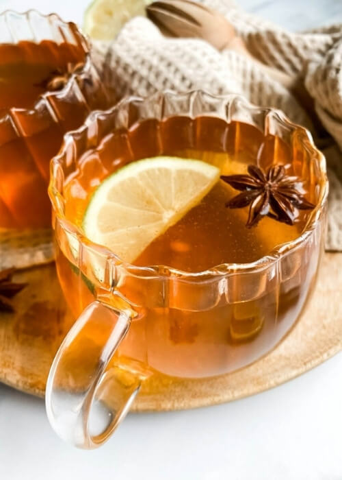 Festive Hot Toddy Mocktail Recipe, Warm & Delicious 3 close up, two cups of hot toddy mocktail with slices of lemon and star anise, over a wooden board and light brown tea towel, slices of lemons, star anise and honey around, hand holding one cup