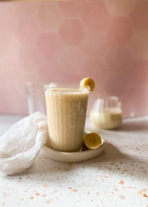 Easy Korean Banana Milk Recipe - Super Creamy 바나나 우유 4 banana milk in a glass, on a tray with sliced banana, a towel, another glass and jug behind, a pink and speckled background