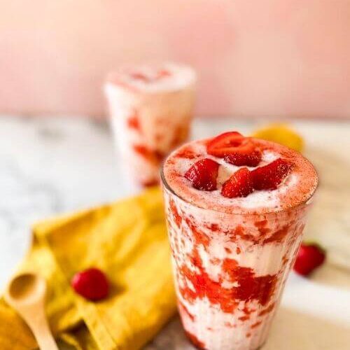 from 45 angle - two glasses of korean strawberry m, on the side a yellow towel, a spoon and some strawberries, on a pink and marble background