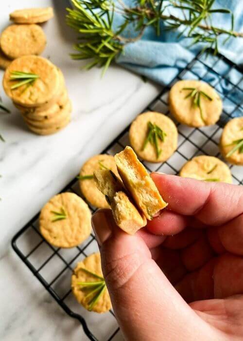 Most Delicious Savory Shortbread With Parmesan And Rosemary, 3 savory shortbread cookies with rosemary on top, on a cooling rack, under a blue towel, more cookies around, on a marble background, hand holding two halves of a cookie