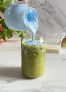 Easy Spirulina Matcha Latte Drink Recipe 12 spirulina matcha latte being made, spirulina cold foam being poured, a plant, a milk jug and chansen in the background, on a marble background