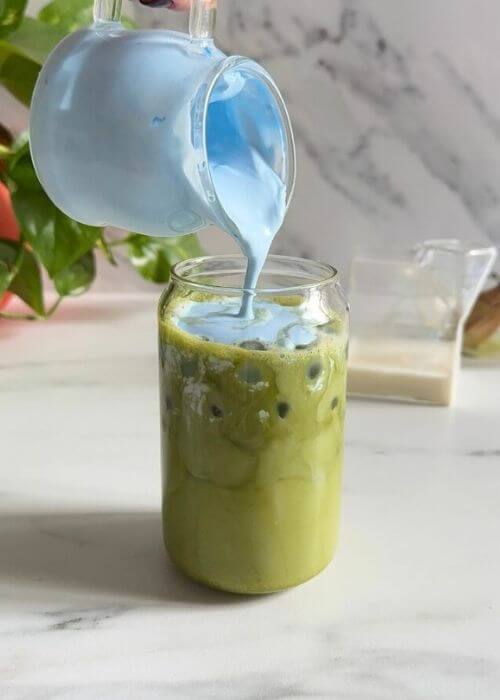 Easy Spirulina Matcha Latte Drink Recipe 5 spirulina matcha latte being made, spirulina cold foam being poured, a plant, a milk jug and chansen in the background, on a marble background