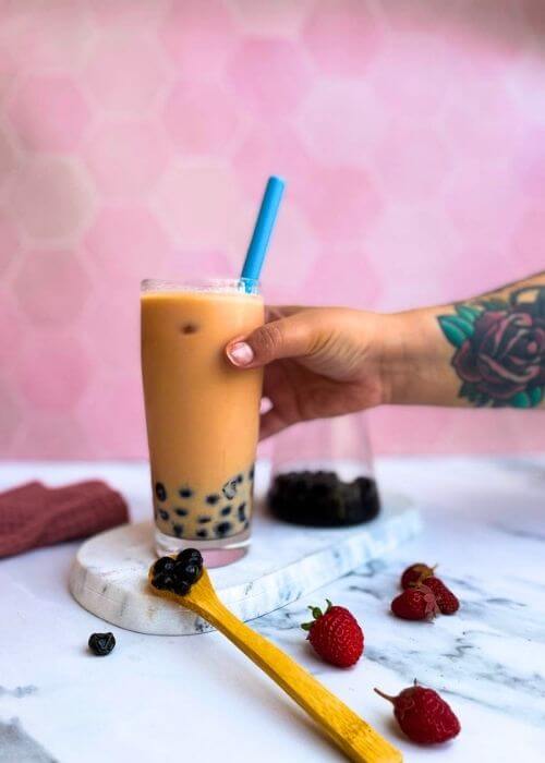 a strawberry milk tea on a marble tray with a glass of boba on pink background, a hand on the glass