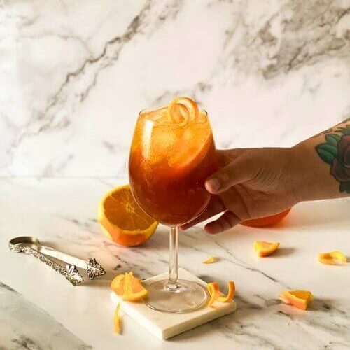 Super Aperol Spritz Mocktail, Just Like The Real One 4 hand reaching for aperol spritz mocktail, marble background, ice tongues and slices of orange around