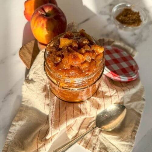apples caramelized in a jar, over a striped towel, a spoon, apples and brown sugar on the sides, over a marble background