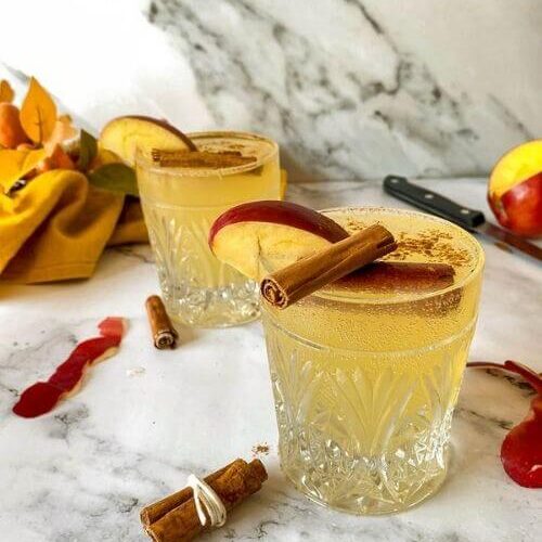 Quick Apple Fizz Mocktail, Delicious Fall Drink 4 apple fizz mocktail with apple slices and cinnamon sticks on top, apple peels, cinnamon sticks, knife, towel and leaves around, on marble background, glass bottle of ginger beer