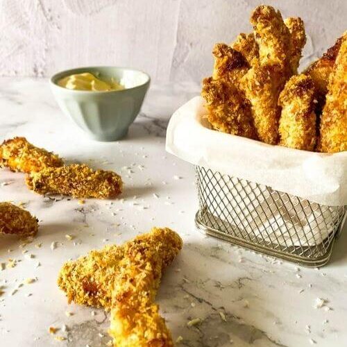 chicken fries in a container with parchment paper on a marble background, more fries around and panko, behind a bowl with mayo