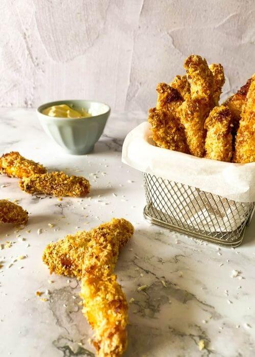 chicken fries in a container with parchment paper on a marble background, more fries around and panko, behind a bowl with mayo