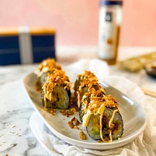 Easy & Quick Chicken Tempura Roll Recipe 4 chicken tempura roll on a plate, under a towel, in the background a bento box, crispy onions and chopsticks, on marble table and pink background