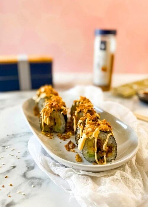 chicken tempura roll on a plate, under a towel, in the background a bento box, crispy onions and chopsticks, on marble table and pink background