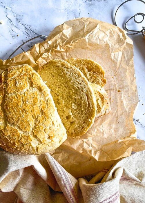 3 Minutes No Knead Bread Without Dutch Oven 6 no knead bread, under parchment paper, wire rack, towel and on the side a bread a dough whisk, on marble background, slices pn the paper