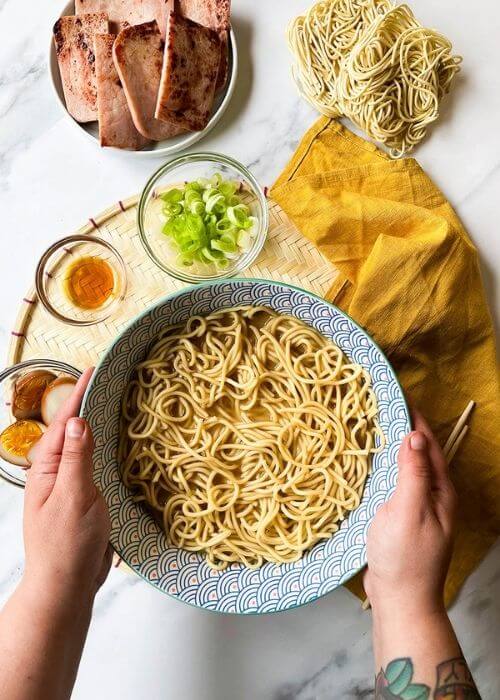 The Ponyo Ramen Recipe You Can't Miss Out On Because It's Delicious 3 two hands holding a bowl of noodles, ingredients around in bowls and towel in the back, on a marble background - from top