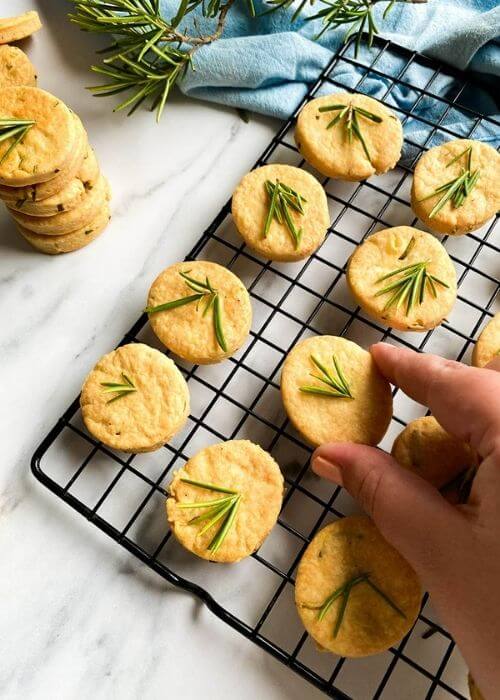 Most Delicious Savory Shortbread With Parmesan And Rosemary, 2 savory shortbread cookies with rosemary on top, on a cooling rack, under a blue towel, more cookies around, on a marble background, hand holding a cookie
