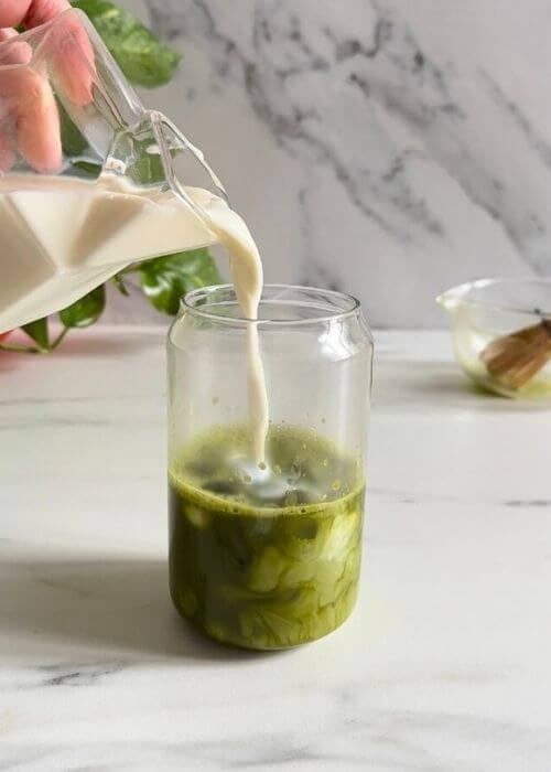 Easy Spirulina Matcha Latte Drink Recipe 4 spirulina matcha latte being made, milk being poured in the glass, plant and matcha bowl with chansen behind