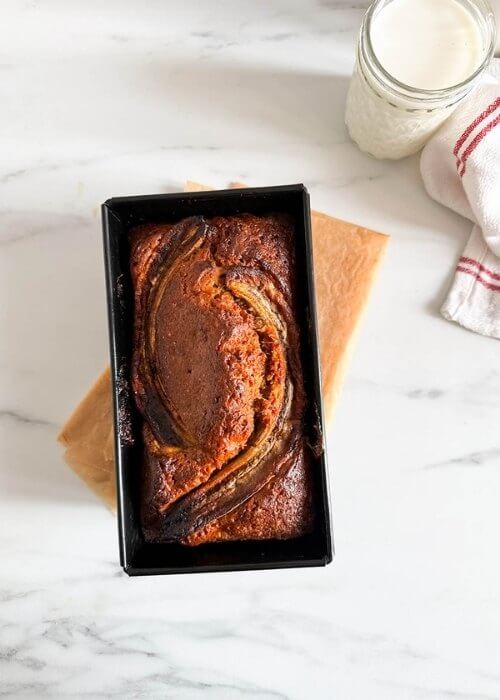 4 ingredient banana bread in a loafpan, on marble background, a glass of milk and towel on the side