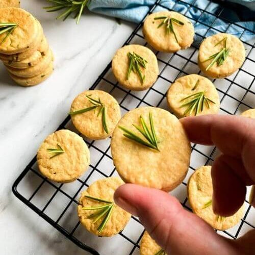 Most Delicious Savory Shortbread With Parmesan And Rosemary, 7 close up - savory shortbread cookies with rosemary on top, on a cooling rack, under a blue towel, more cookies around, on a marble background, a hand holding a cookie