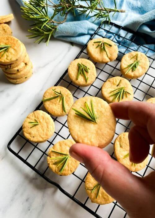close up - savory shortbread cookies with rosemary on top, on a cooling rack, under a blue towel, more cookies around, on a marble background, a hand holding a cookie