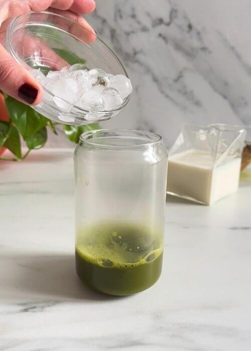 Easy Spirulina Matcha Latte Drink Recipe 3 spirulina matcha latte being made, ice being poured in the glass, plant and matcha bowl with chansen behind, jug of milk