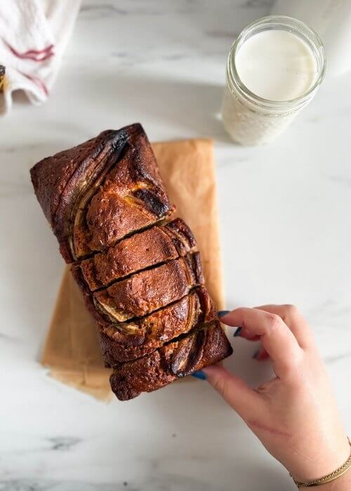 4 ingredient banana bread in a loafpan, on marble background, a glass of milk and towel on the side, hand reaching for a slice