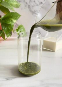 Easy Spirulina Matcha Latte Drink Recipe 10 spirulina matcha latte being made, matcha being poured in the glass, plant and jug of milk behind