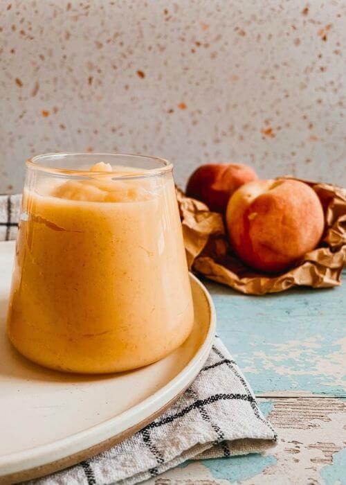 peach curd in a jar on a plate, under a towel, in the back two peaches on a paper bag