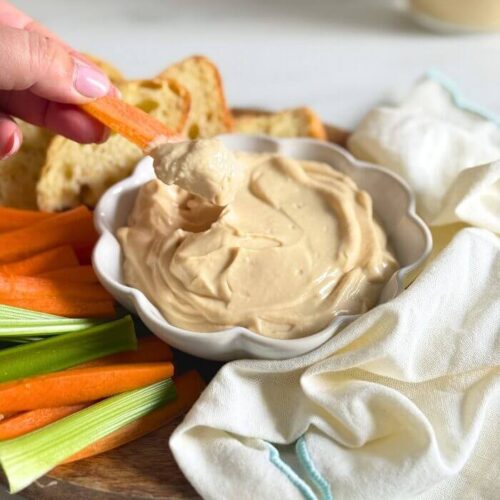 hand holding a carrot dipping into a bowl of miso mayonnaise, on a wooden board with more veggies cut on the side, slices of bread, towel and a jar of mayo in the back