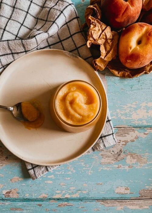 The Most Delicious Peach Curd Recipe 2 from top peach curd in a jar on a plate with a spoon, under a towel, in the back two peaches on a paper bag
