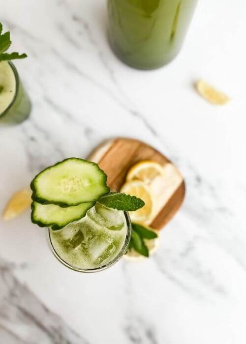cucumber mint lemonade in a glass on a wooden tray, around slices of lemon, and more cucumber lemonade, on marble background - from top
