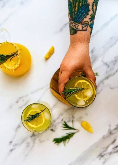 hand holding a glass of limoncello tonic on a wooden coaster, a jug of lemon juice with lemon slices and rosemary on the side, another glass of limoncello on. a marble background - from top