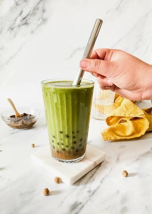 Easy Boba Smoothie Recipe With Banana And Coconut Milk 3 hand holding a straw in a glass of boba smoothie with boba pearls around and in a bowl with a spoon, in the back two empty glasses and a yellow towel, on marble background