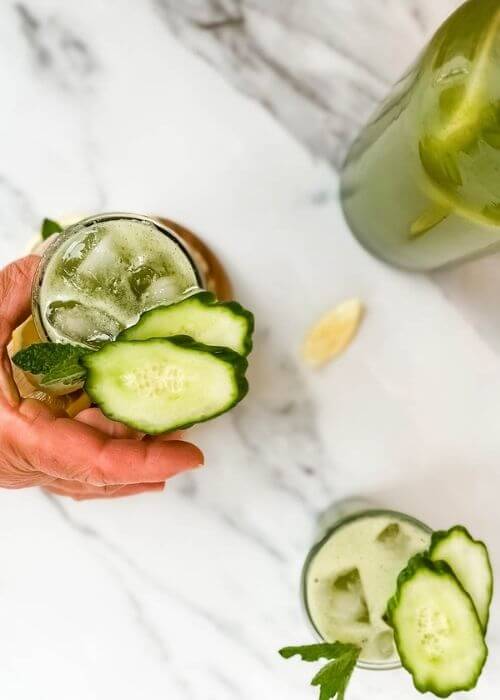 hand reaching for one glass of cucumber mint lemonade in a glass on a wooden tray, around slices of lemon, and more cucumber lemonade, on marble background - from top