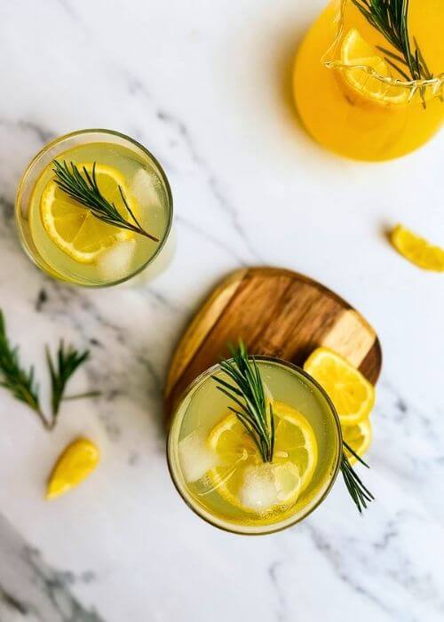 two glass of limoncello tonic on a wooden coaster, a jug of lemon juice with lemon slices and rosemary on the side,a marble background