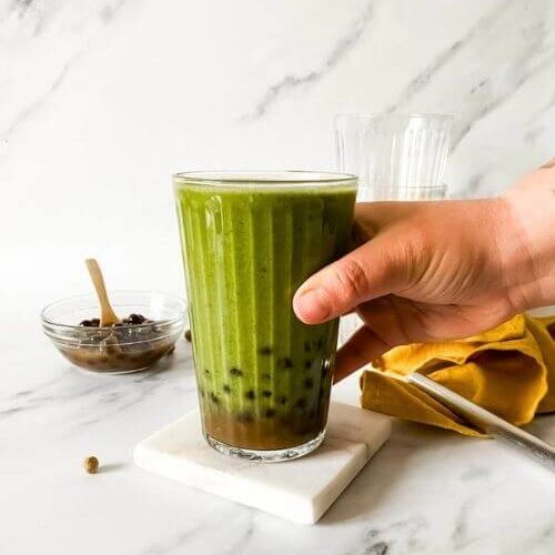 hand holding a glass of boba smoothie with boba pearls around and in a bowl with a spoon, in the back two empty glasses and a yellow towel, on marble background