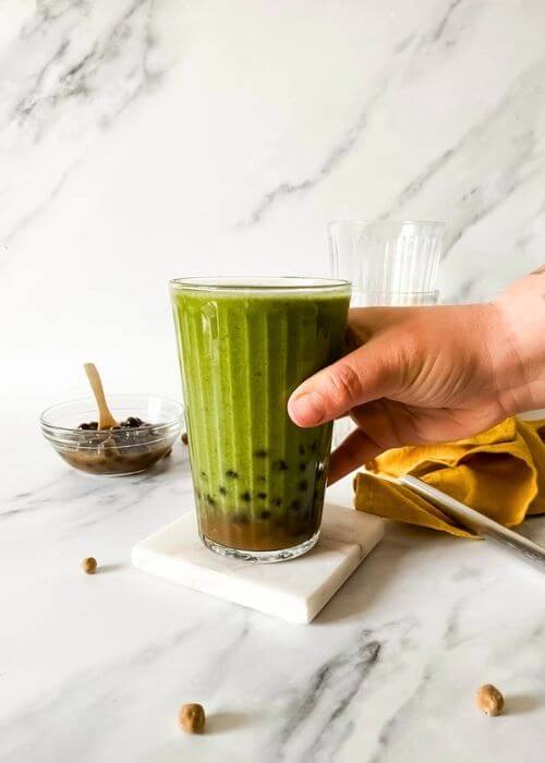 Easy Boba Smoothie Recipe With Banana And Coconut Milk 4 hand holding a glass of boba smoothie with boba pearls around and in a bowl with a spoon, in the back two empty glasses and a yellow towel, on marble background