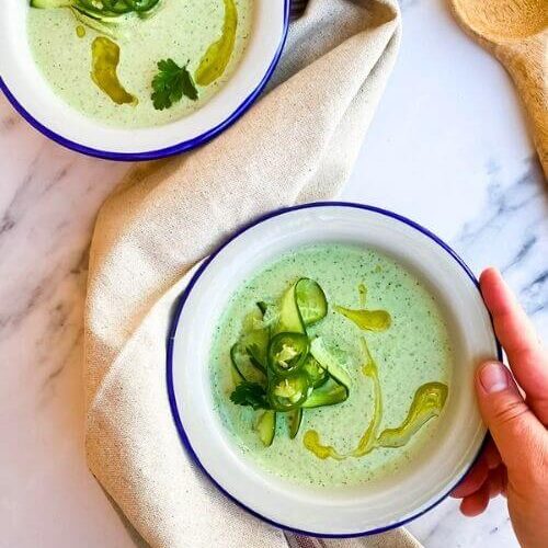 from top - two bowls of cucumber gazpacho on a towel, spoon on the side, on marble background, hands holding one bowl