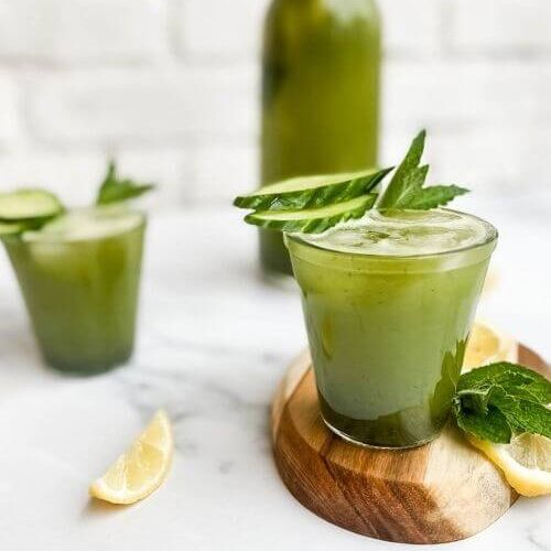 from front - two glasses of cucumber mint lemonade, one on a wooden tray, slices of lemon and mint leaves around, in the back a jug of lemonade on marble background