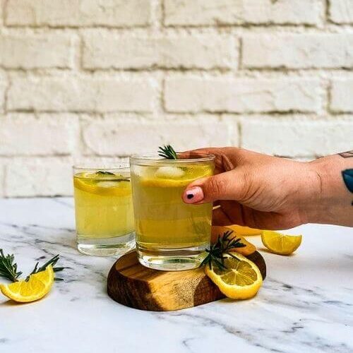 hand holding a glass of limoncello tonic on a wooden coaster, a jug of lemon juice with lemon slices and rosemary on the side, another glass of limoncello on. a marble background - from frony
