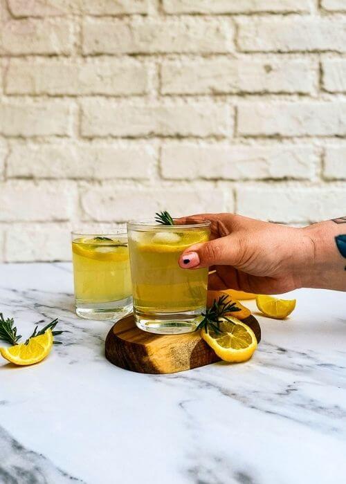 hand holding a glass of limoncello tonic on a wooden coaster, a jug of lemon juice with lemon slices and rosemary on the side, another glass of limoncello on. a marble background - from frony