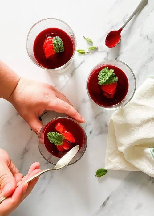 The Best Vegan Panna Cotta 5 hand holding a glass of vegan panna cotta in glasses on a marble background, a spoon and mint leaves on the side, towel on the side
