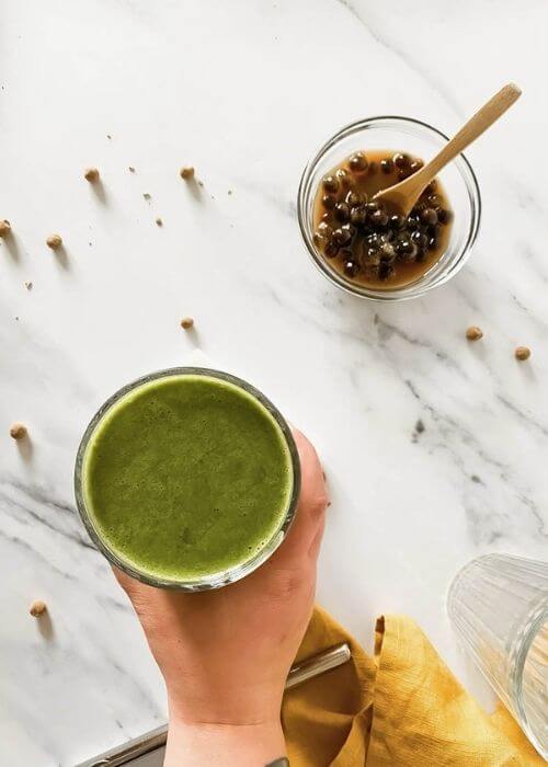Easy Boba Smoothie Recipe With Banana And Coconut Milk 2 from top - hand holding a glass of boba smoothie with boba pearls around and in a bowl with a spoon, in the back two empty glasses and a yellow towel, on marble background