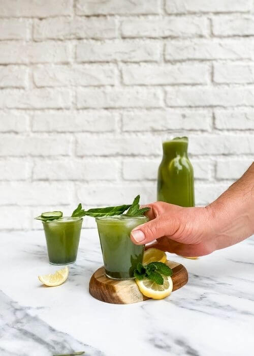 from front - two glasses of cucumber mint lemonade, hand holding one of them, one on a wooden tray, slices of lemon and mint leaves around, in the back a jug of lemonade on marble background