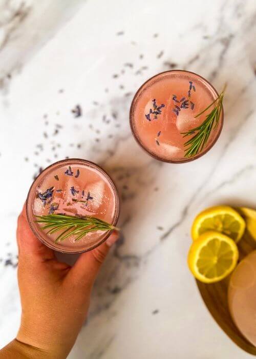 Lavender Lemonade Cocktail 20 Minutes Delicious Drink 2 hand reaching in for glass of lavender cocktail, jug with cocktail mix and lemons, marble background
