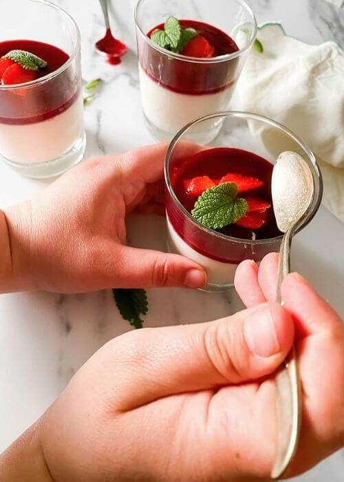 The Best Vegan Panna Cotta 3 close up of hands holding a vegan panna cotta in glasses on a marble background, a spoon and mint leaves on the side, towel on the side