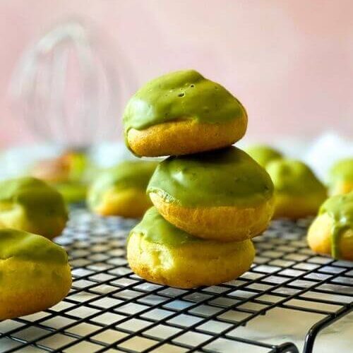 stack of matcha cream puffs on a wire rack, behind a whisk, a white towel on a pink tiled background