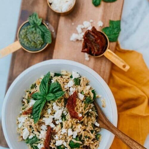 orzo pesto salad in a bowl with a spoon, pesto, sundried tomatoes and feta in measuring cups, on a wooden board, around a yellow towel