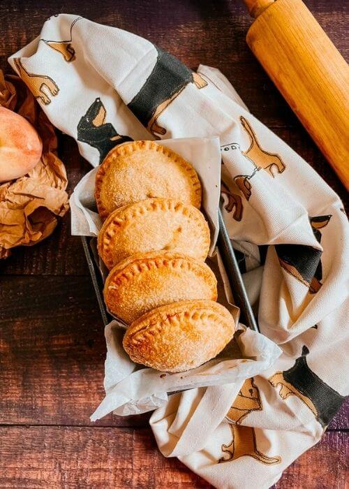 Super Easy Peach Hand Pies With a Fresh Basil Filling 4 peach hand pies in a tin, a napkin under, rolling pin and peaches on the sides