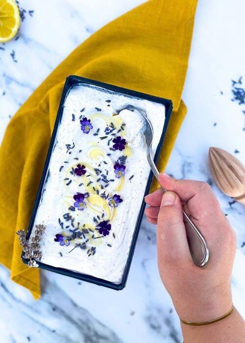 The Best Lavender Lemon Ice Cream Recipe To Try Immediately 4 from top - hand with a spoon spooning some lavender lemon ice cream in a loaf pan, a yellow towel under, lemon, lemon squeezer and lavender buds around, on a marble background