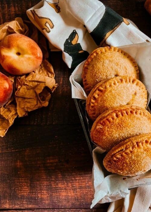 Super Easy Peach Hand Pies With a Fresh Basil Filling 3 peach hand pies from above, a napkin under them and peaches on the side